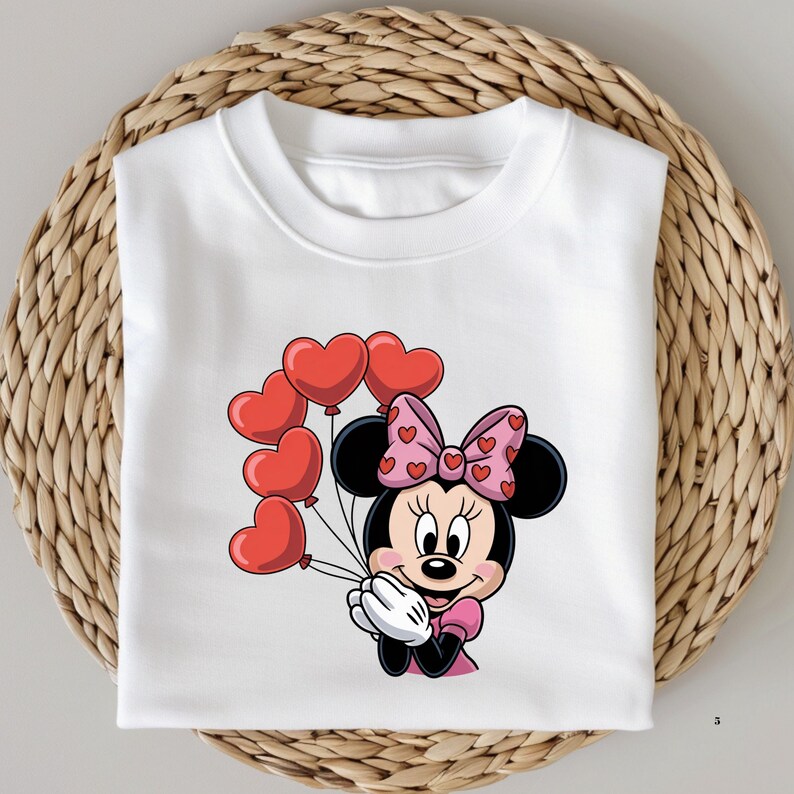 May include: White sweatshirt featuring a cartoon Minnie Mouse holding red heart-shaped balloons. Minnie Mouse is wearing a pink dress and a pink bow with heart patterns. The sweatshirt is folded on a woven surface.