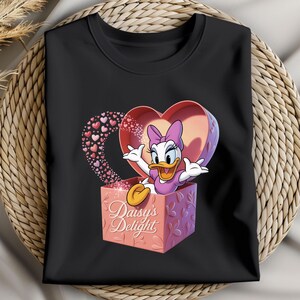 May include: Black crew neck t-shirt featuring a cartoon image of Daisy Duck emerging from a heart-shaped box. The box is pink and has the words "Daisy's Delight" printed on it. A trail of small hearts extends from the box.