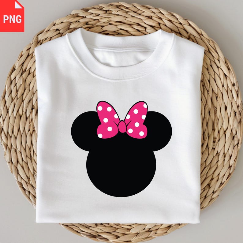 K&ouml;nnte beinhalten: Wei&szlig;es Sweatshirt mit Rundhalsausschnitt und schwarzer Maus-Kopf-Silhouette und rosa Schleife. Die Schleife hat wei&szlig;e Tupfen. Das Sweatshirt ist auf einer gewebten, hellbraunen Oberfl&auml;che gefaltet.