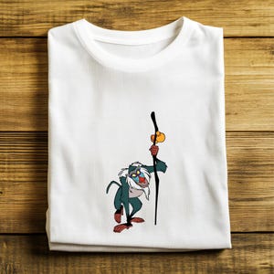 May include: White t-shirt featuring a cartoon illustration of a monkey-like character holding a staff. The character is depicted in shades of green, blue, red, and yellow. The t-shirt is folded and placed on a wooden surface.