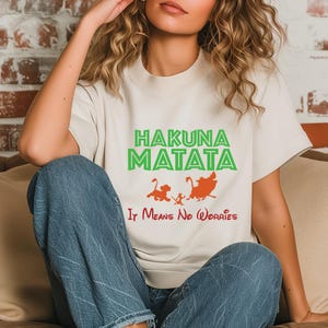 May include: Cream-colored t-shirt with the words "HAKUNA MATATA" in green and "It Means No Worries" in red. Features cartoon illustrations of Simba, Timon, and Pumbaa in orange. A casual, comfortable tee.