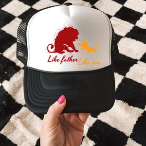 May include: Black and white trucker hat with a white front panel featuring a red lion and a yellow cub graphic. The text "Like father Like son" is printed below the graphic in red and yellow.