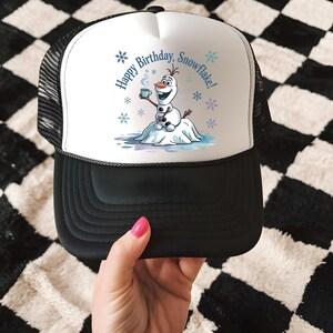 May include: A black and white trucker hat with a white front panel featuring a cartoon snowman holding a mug. The text "Happy Birthday, Snowflake!" is printed above the snowman, with snowflake graphics.