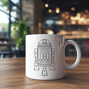 May include: White ceramic mug featuring a black line drawing of the Star Wars character R2-D2. The mug has a rounded handle and is set on a wooden table.