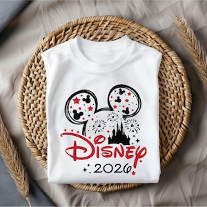 May include: White t-shirt featuring a black and red Mickey Mouse head design with stars and fireworks. The word "Disney" is in red script, with "2026" below. The shirt is sleeveless and folded on a woven basket.