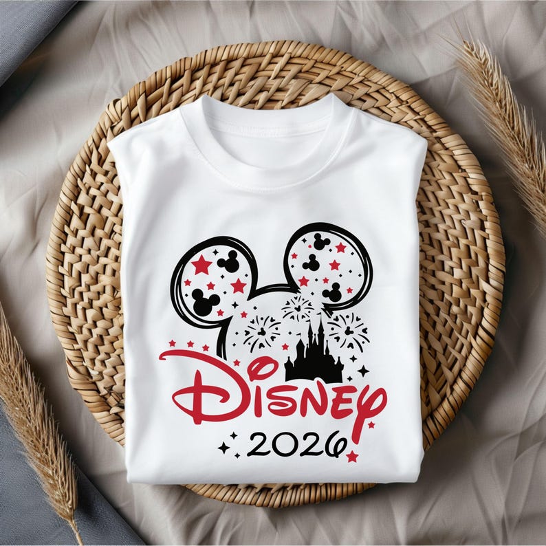 May include: White t-shirt featuring a black and red Mickey Mouse head design with stars and fireworks. The word "Disney" is in red script, with "2026" below. The shirt is sleeveless and folded on a woven basket.