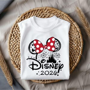 May include: White t-shirt featuring a Minnie Mouse bow with red polka dots, black Mickey Mouse ears, and a silhouette of a castle. The word "Disney" and the year "2026" are printed in black.