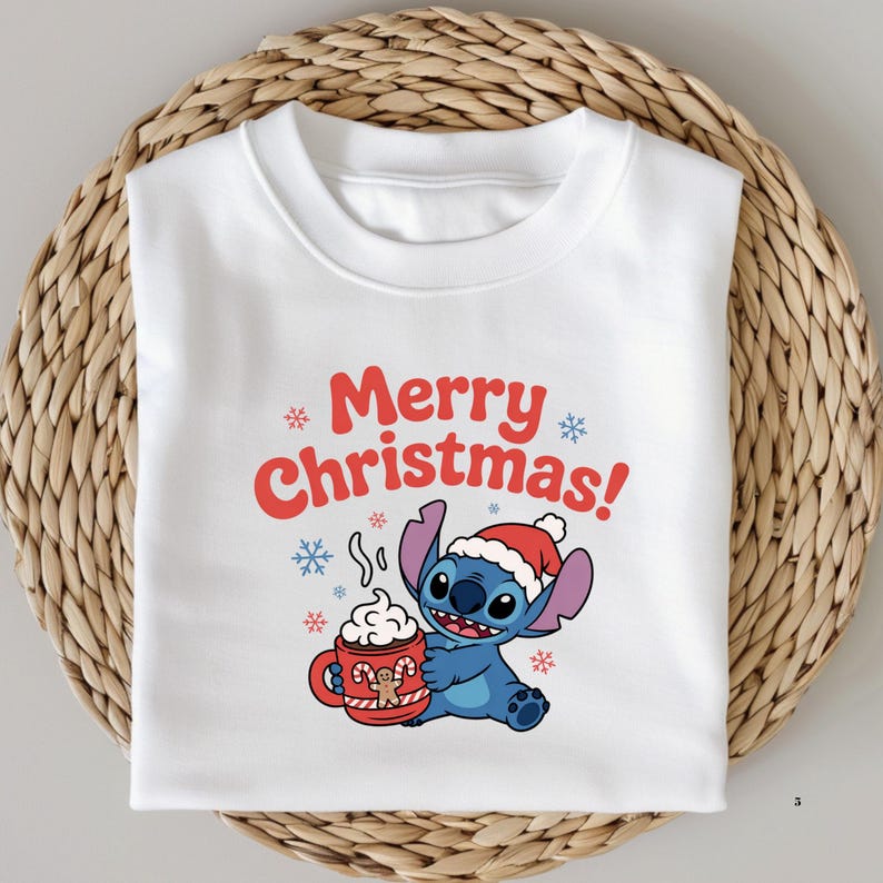 Pu&ograve; includere: Felpa bianca con un personaggio dei cartoni animati di Stitch con un cappello da Babbo Natale e una tazza di cioccolata calda. Il testo "Merry Christmas!" &egrave; in rosso con fiocchi di neve. La felpa &egrave; piegata su una superficie tessuta.