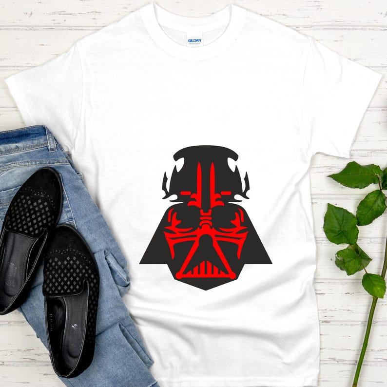 May include: White t-shirt featuring a black and red graphic of a character's helmet. The graphic is centered on the shirt. The shirt is paired with blue jeans and black slip-on shoes. The shirt is made of a soft material.