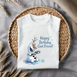 May include: White t-shirt featuring a cartoon snowman holding a mug, with the text "Happy Birthday, Cool Friend!" in blue. The snowman has a carrot nose and twig hair. The shirt is displayed on a woven basket.