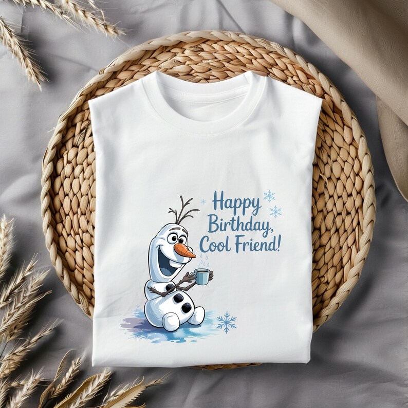 May include: White t-shirt featuring a cartoon snowman holding a mug, with the text "Happy Birthday, Cool Friend!" in blue. The snowman has a carrot nose and twig hair. The shirt is displayed on a woven basket.