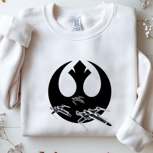 May include: White crewneck sweatshirt featuring a black graphic of the Rebel Alliance symbol with three X-wing starfighters. The design is centered on the front of the long-sleeved top.
