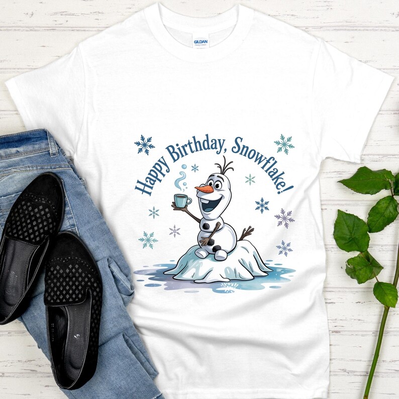 May include: White t-shirt featuring a cartoon snowman holding a mug with the text "Happy Birthday, Snowflake!" in blue. The design includes snowflakes. The shirt is paired with blue jeans and black slip-on shoes.