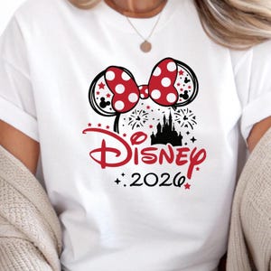May include: White t-shirt featuring a red and white polka dot bow, black Mickey Mouse ears, and a black silhouette of a castle. The word "Disney" is in red script, with "2026" below. Red stars and fireworks complete the design.