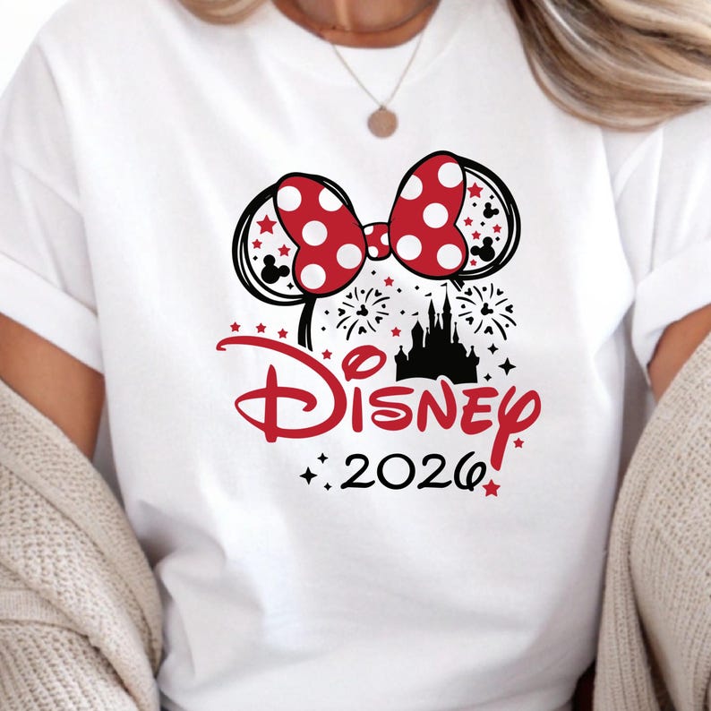 May include: White t-shirt featuring a red and white polka dot bow, black Mickey Mouse ears, and a black silhouette of a castle. The word "Disney" is in red script, with "2026" below. Red stars and fireworks complete the design.