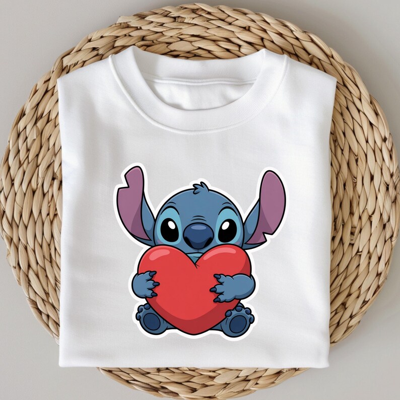 May include: White t-shirt featuring a cartoon illustration of Stitch, a blue alien character, holding a large red heart. The design is centered on the shirt, showcasing the character's large eyes and ears. The shirt is folded on a woven surface.