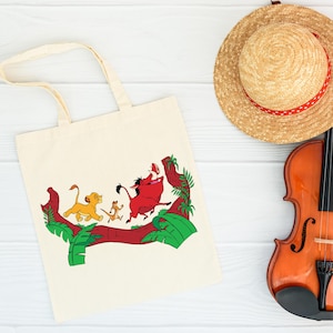 May include: Cream-colored tote bag featuring a colorful graphic of characters from The Lion King. The design includes Simba, Timon, Pumbaa, and a red log with green foliage. A straw hat with a red ribbon and a violin are also visible.