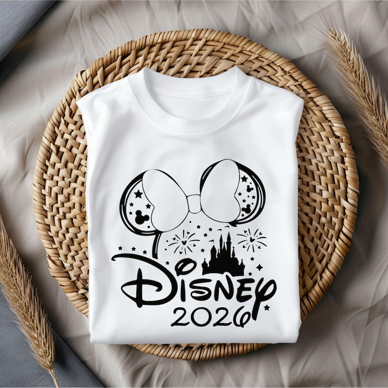May include: White t-shirt featuring a black graphic of Minnie Mouse ears with stars, a bow, fireworks, and a castle. The word "Disney" and the year "2026" are printed below the graphic. The shirt is displayed on a woven basket.