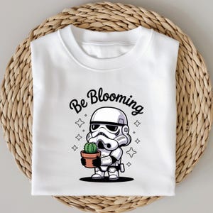 May include: White sweatshirt featuring a cartoon Stormtrooper holding a potted cactus. The phrase "Be Blooming" is written above the character in black script. The design includes star accents, creating a playful and whimsical aesthetic.