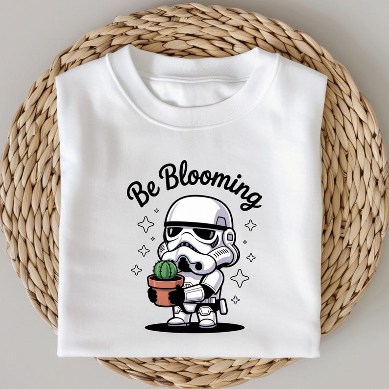 May include: White sweatshirt featuring a cartoon Stormtrooper holding a potted cactus. The phrase "Be Blooming" is written above the character in black script. The design includes star accents, creating a playful and whimsical aesthetic.