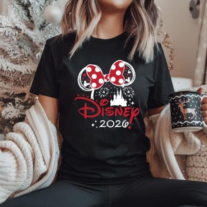 May include: Black t-shirt featuring a red and white polka dot bow, Mickey Mouse ears, and the word "Disney" in red script. The design includes a castle, fireworks, and the year "2026".