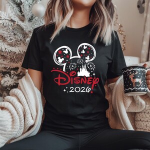 May include: Black t-shirt featuring a Disney design with Mickey Mouse ears, fireworks, and a castle graphic. The text "Disney 2026" is in red. A festive mug with a deer design is also visible.