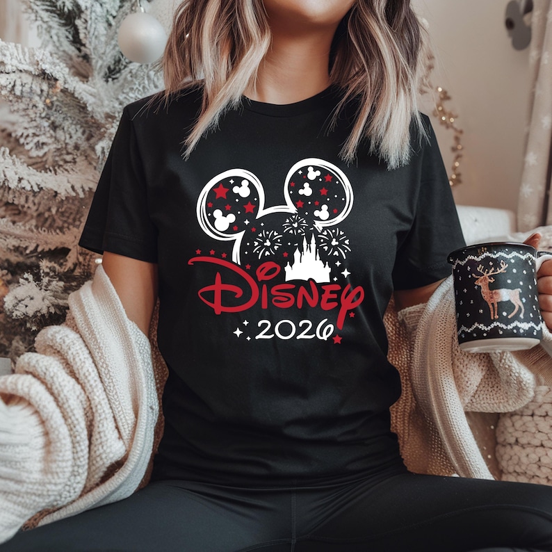 May include: Black t-shirt featuring a Disney design with Mickey Mouse ears, fireworks, and a castle graphic. The text "Disney 2026" is in red. A festive mug with a deer design is also visible.