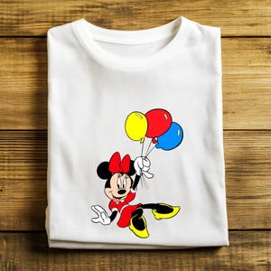May include: White t-shirt featuring a cartoon Minnie Mouse character in a red dress and bow, holding yellow, red, and blue balloons. The shirt is folded on a wooden surface.