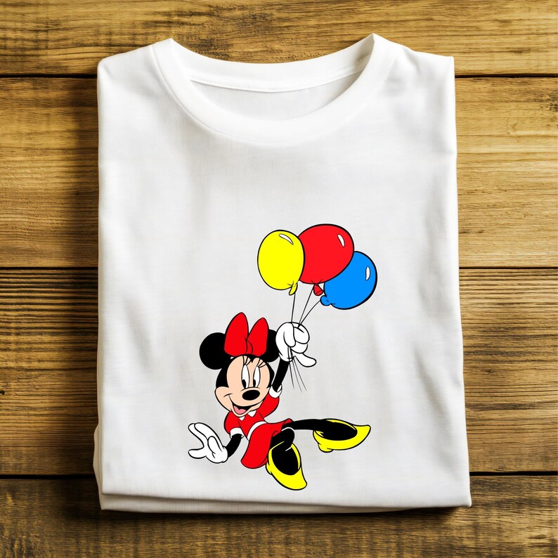 May include: White t-shirt featuring a cartoon Minnie Mouse character in a red dress and bow, holding yellow, red, and blue balloons. The shirt is folded on a wooden surface.