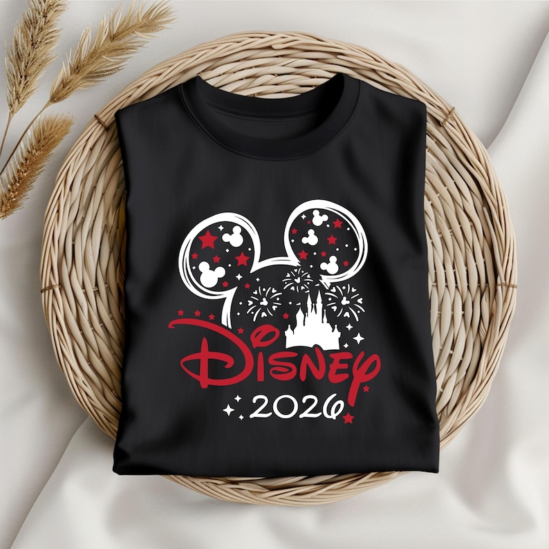 May include: Black t-shirt featuring a Disney design. The design includes Mickey Mouse ears filled with red stars and white Mickey heads, fireworks, a castle, and the word "Disney" in red with the year "2026".