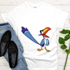 May include: White t-shirt featuring a cartoon bird character with a long blue tail, red beak, and yellow feet. The shirt is paired with blue jeans and black slip-on shoes. The shirt is made of a soft material.