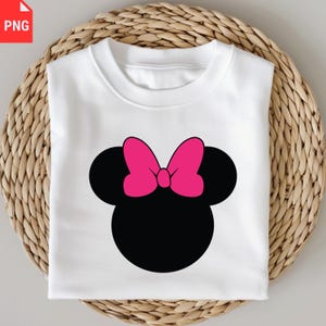 K&ouml;nnte beinhalten: Wei&szlig;es Sweatshirt mit Rundhalsausschnitt und schwarzer Maus-Kopf-Silhouette mit leuchtend rosa Schleife. Das Sweatshirt liegt gefaltet auf einer gewebten, hellbraunen Oberfl&auml;che. Das Design ist mittig auf der Vorderseite des Kleidungsst&uuml;cks platziert.