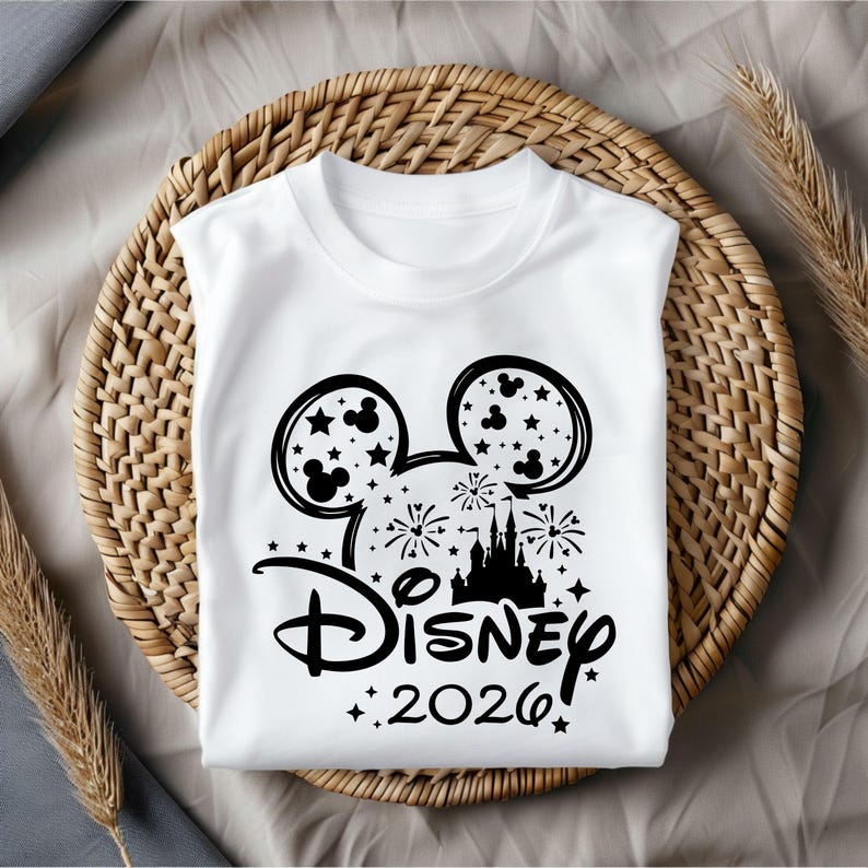 May include: White t-shirt featuring a black Mickey Mouse design with stars, fireworks, and a castle silhouette. The word "Disney" is written in a stylized font, with "2026" below. The shirt is displayed on a woven basket.