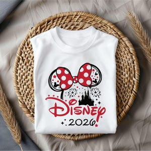 May include: White sleeveless t-shirt featuring a Minnie Mouse bow with red polka dots and black mouse ears. The shirt has a Disney castle graphic, fireworks, and the text "Disney 2026" in red.