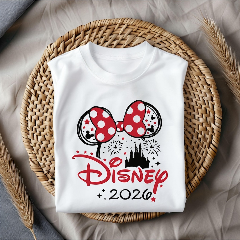 May include: White sleeveless t-shirt featuring a Minnie Mouse bow with red polka dots and black mouse ears. The shirt has a Disney castle graphic, fireworks, and the text "Disney 2026" in red.