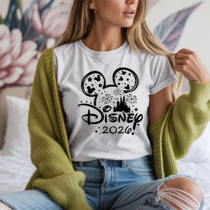May include: White t-shirt featuring a black graphic of Mickey Mouse ears with stars, fireworks, and a castle. The word "Disney" is written in a stylized font, with "2026" below. The person is wearing a green cardigan and blue jeans.