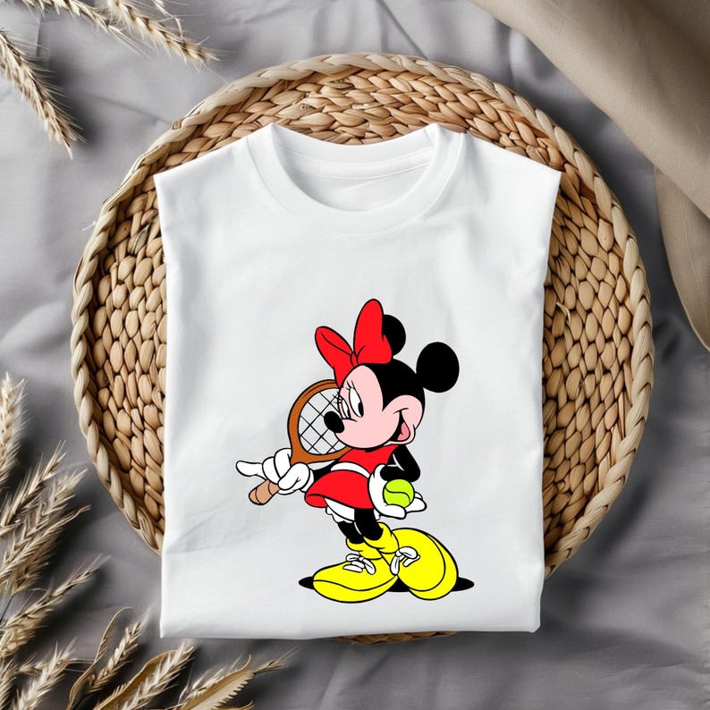 May include: White t-shirt featuring a cartoon Minnie Mouse in a red dress and bow, holding a tennis racket and ball. The design includes yellow shoes and is set against a woven basket background.