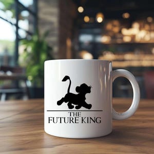 May include: White ceramic mug featuring a black silhouette of a lion cub above the text "THE FUTURE KING". The mug has a curved handle and is set on a wooden surface. The design is simple and bold.