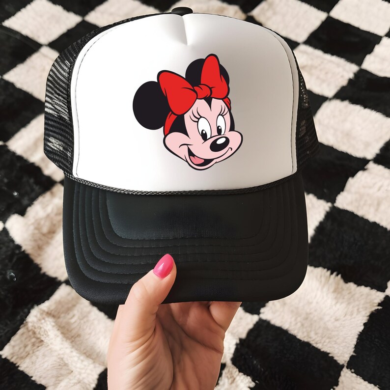 May include: A black and white trucker hat with a white front panel featuring a Minnie Mouse graphic. The graphic shows Minnie Mouse's face with a red bow. The hat has a black brim and a mesh back.