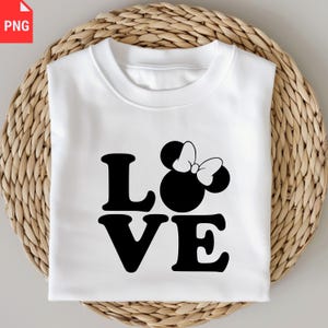 May include: White sweatshirt with the word "LOVE" in bold black letters. The "O" is replaced with a Minnie Mouse head and bow design. The sweatshirt is folded and placed on a woven, circular surface.