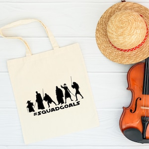 May include: Cream-colored tote bag with a black graphic of stylized figures and the text "#SQUADGOALS." A straw hat with a red ribbon and a violin are also in the image.