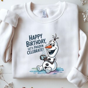 May include: White sweatshirt featuring the text "HAPPY BIRTHDAY, LET'S FROZEN CELEBRATE!" with a cartoon image of Olaf from Frozen. Olaf is holding a cup and sitting on a colorful puddle.