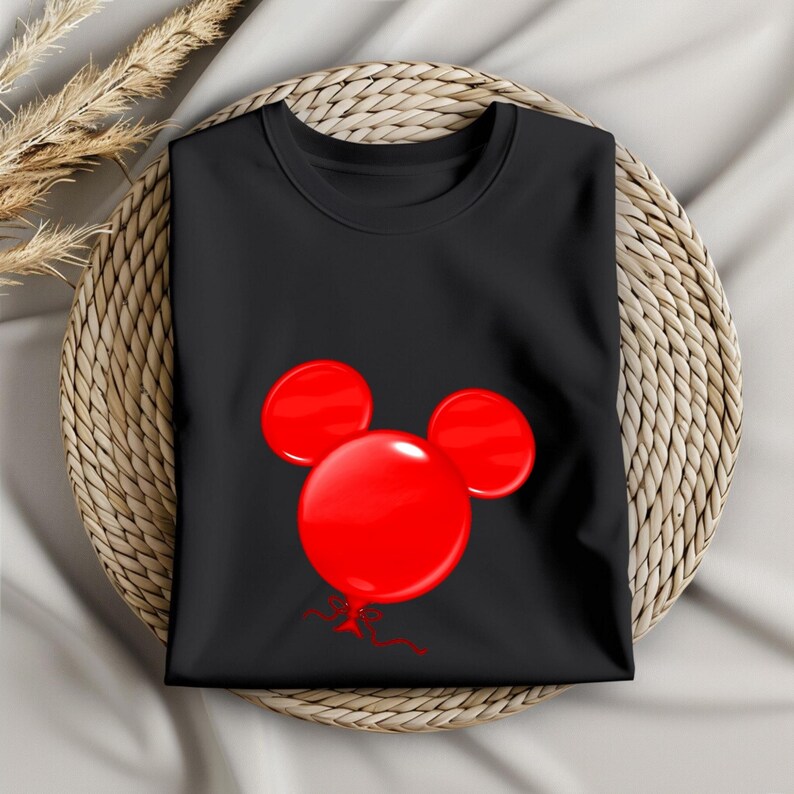 May include: Black t-shirt featuring a red balloon design in the shape of a mouse head. The balloons have a glossy finish and are tied with a red ribbon. The shirt is folded and placed on a woven mat.