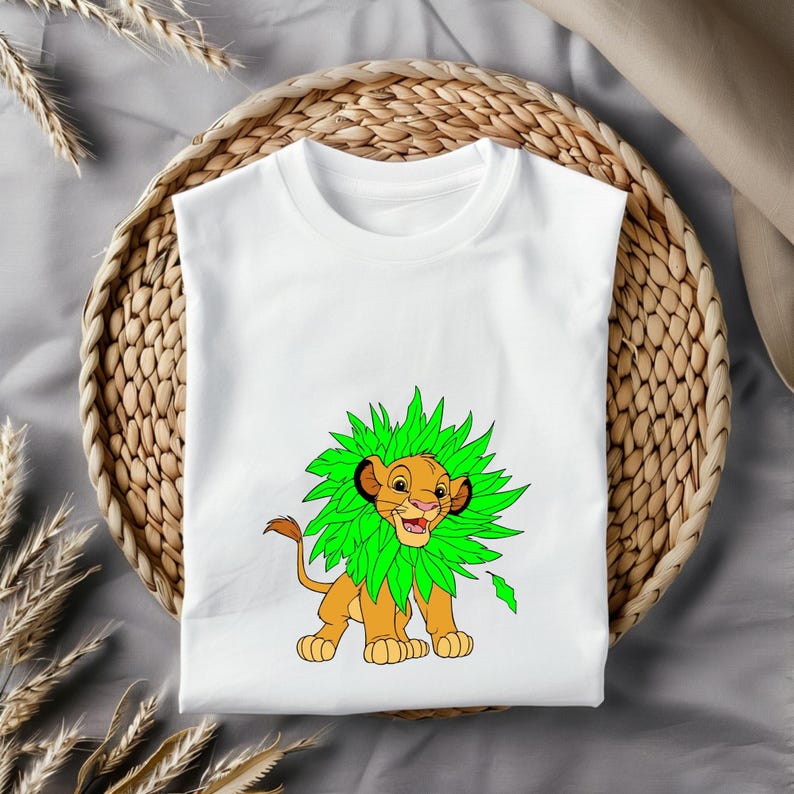 May include: White t-shirt featuring a cartoon lion with a green leafy mane. The lion is depicted in shades of orange and brown, with a smiling expression. The shirt is displayed on a woven basket.