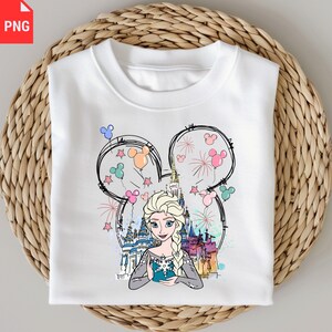 May include: White sweatshirt featuring a graphic of Elsa from Frozen, framed within a Mickey Mouse silhouette. The design includes a castle, fireworks, and colorful balloons. The sweatshirt is folded on a woven surface.
