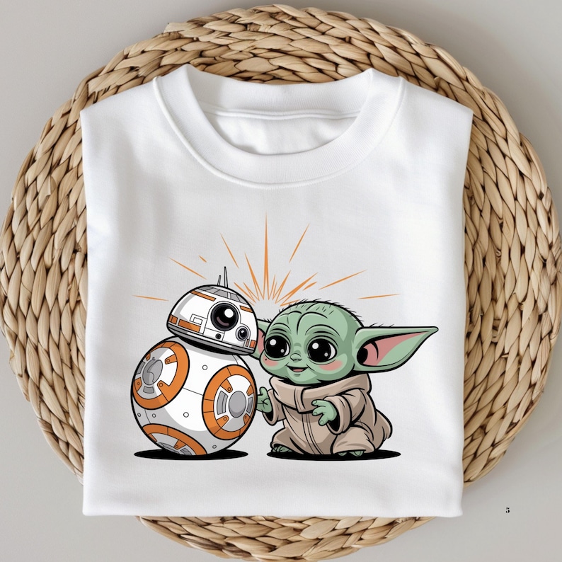 May include: White t-shirt featuring a cartoon illustration of BB-8 and Grogu (Baby Yoda). BB-8 is white and orange, and Grogu is green and wearing a tan robe. The design includes orange starburst lines behind the characters.