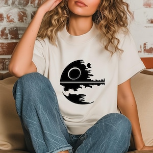 May include: Cream-colored t-shirt featuring a black graphic of a celestial body with a circular design and a detailed lower half. The design is centered on the shirt, offering a bold contrast against the neutral background. The shirt has short sleeves and a crew neck.