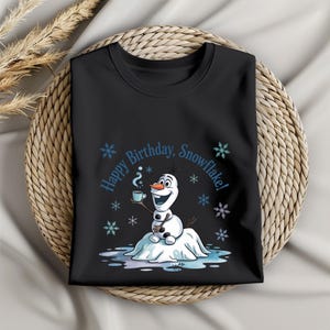 May include: Black t-shirt featuring a cartoon snowman holding a cup of coffee, with the text "Happy Birthday, Snowflake!" surrounded by snowflakes. The snowman is smiling and sitting on a snow mound.