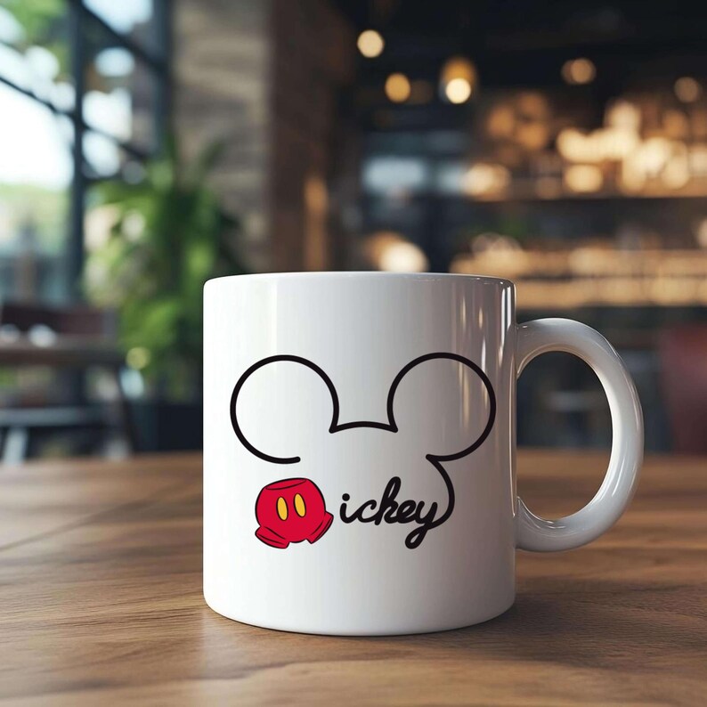 May include: White ceramic mug featuring a black outline of Mickey Mouse's head and ears, with a red and yellow glove and the word "ickey" in a script font. The mug has a white handle and is set on a wooden surface.