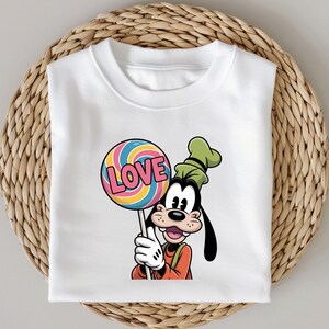 May include: White t-shirt featuring a cartoon image of Goofy holding a colorful lollipop with the word "LOVE". Goofy is wearing an orange shirt and a green hat. The t-shirt is folded on a woven, circular surface.